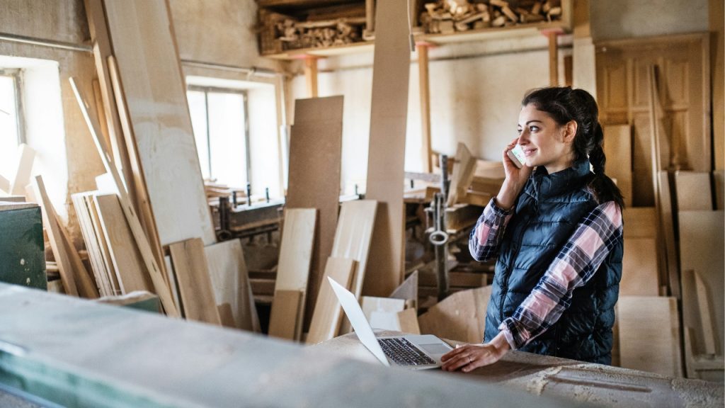 woman-worker-with-smartphone-and-laptop-in-the-carpentry-workshop-making-a-phone-call-